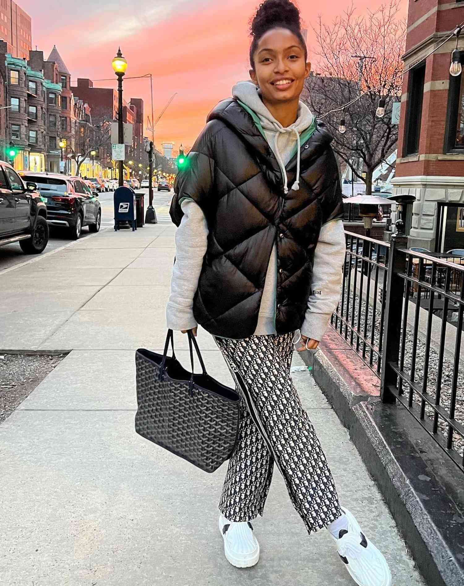 Yara Shahidi wears a black, short-sleeve puffer jacket, a gray hoodie, black and white pants and bag, and white sneakers. 