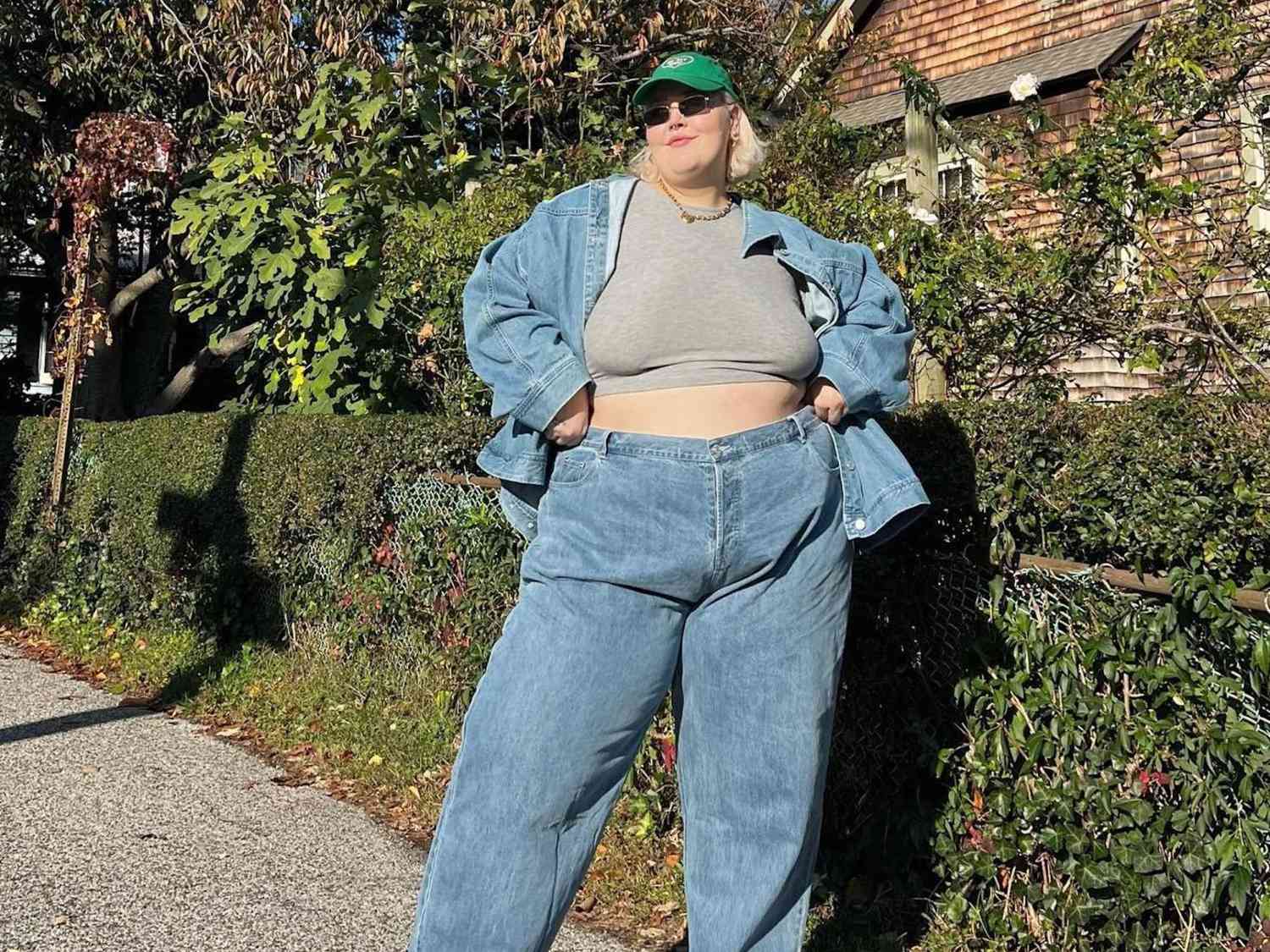 Abby Bible wears a gray cropped t-shirt, denim jacket, jeans, green baseball cap, and sunglasses