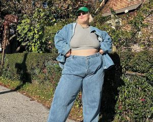 Abby Bible wears a gray cropped t-shirt, denim jacket, jeans, green baseball cap, and sunglasses