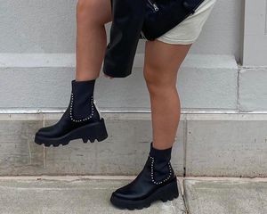 Close-up of woman pulling on studded chunky boots while wearing a leather jacket and shirt dress