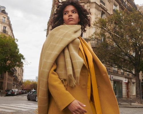 Woman with beige blanket scarf and yellow coat walking through Paris