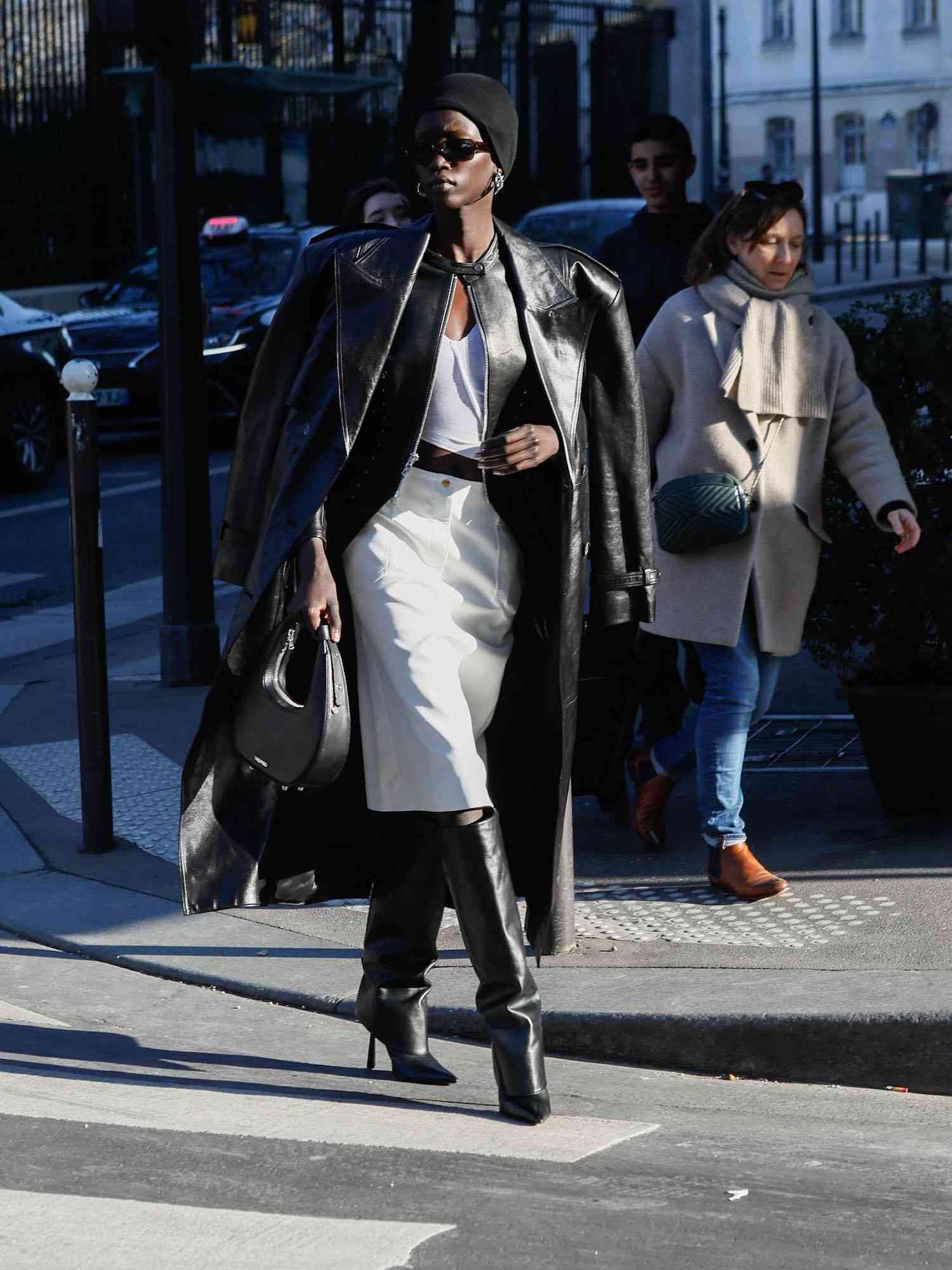 Anok Yai wears a leather trench coat and jacket, white top and midi skirt, knee-high boots, black handbag, beanie, and sunglasses