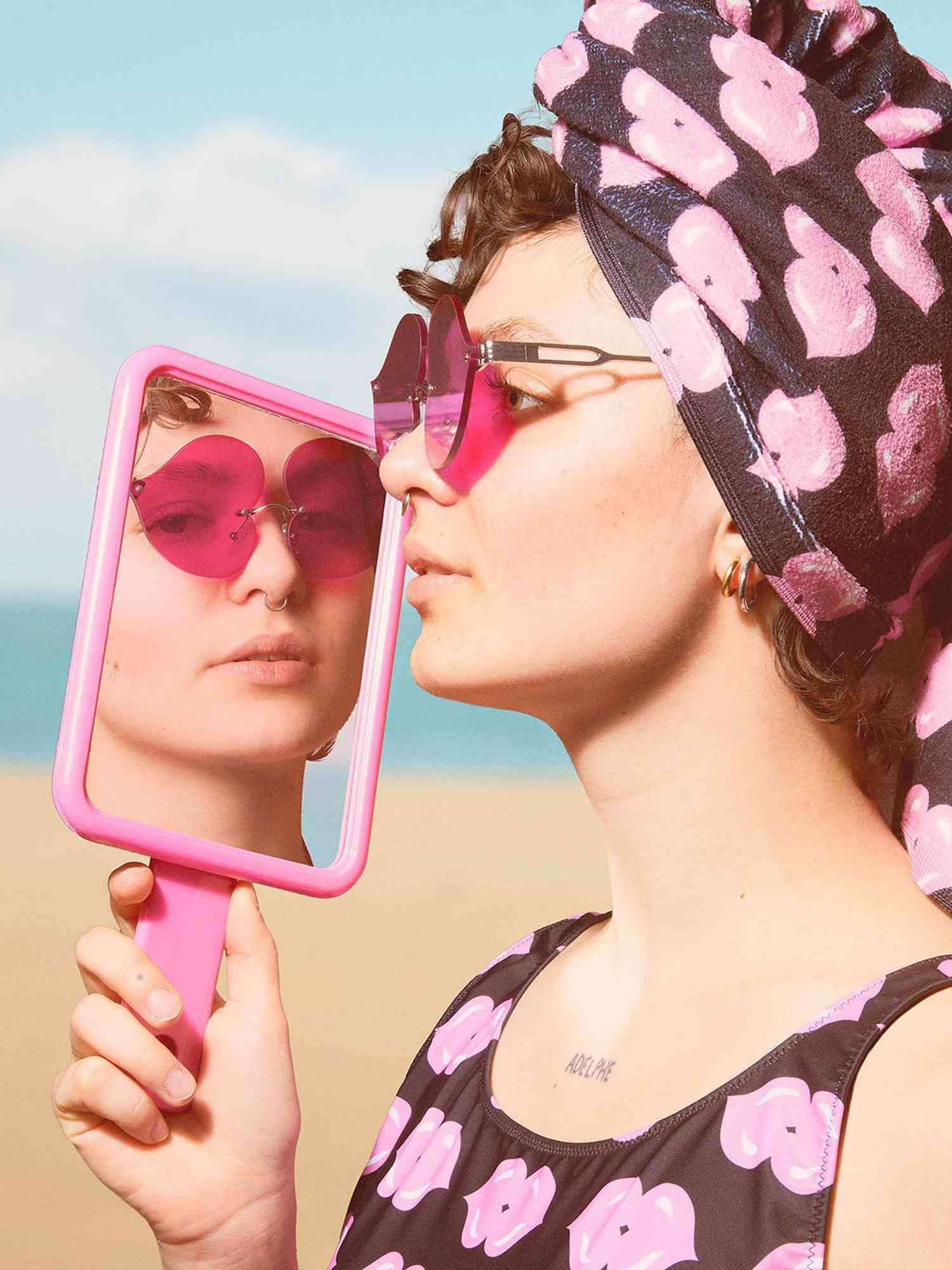 Woman wearing pink kissing lips-patterned towel and swimsuit while holding pink mirror up to face revealing pink sunglasses and septum piercing