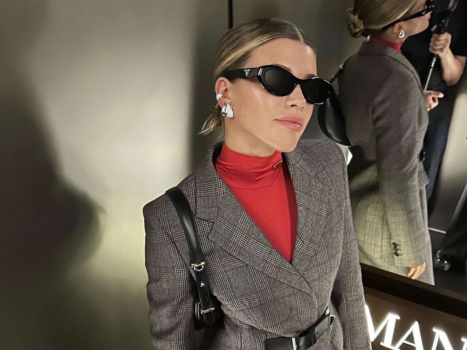 Sofia Richie Grainge wears a red turtleneck, gray houndstooth check coat, black shoulder bag and sunglasses, and silver earrings