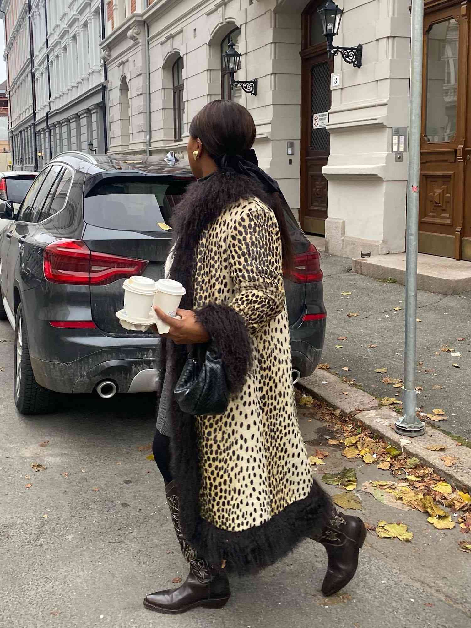 Nnenna Echem wears a leopard print coat with fur trim, cowboy boots, woven handbag, gold earrings, and slicked-back hairstyle while carrying coffee