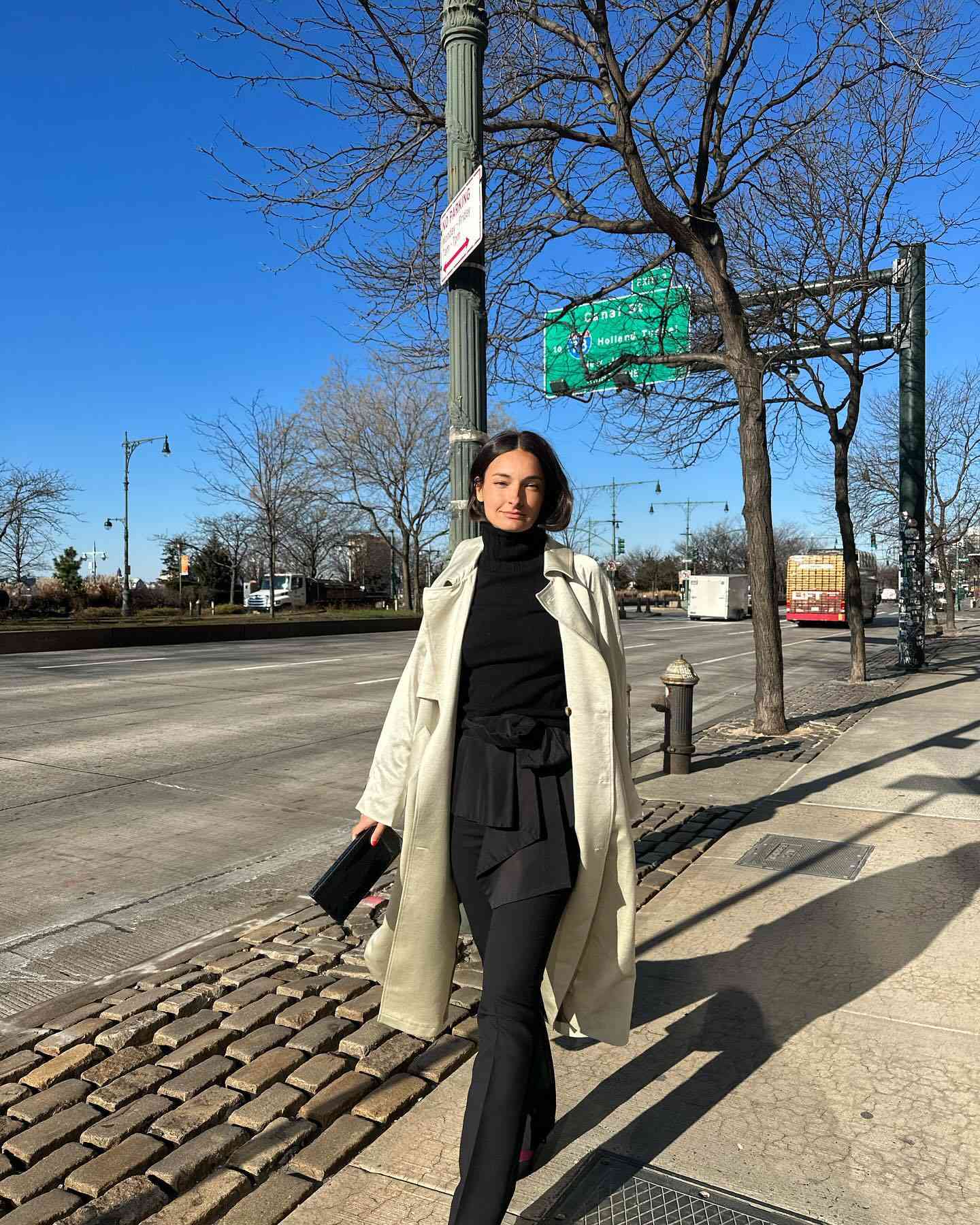 Woman wearing black turtleneck, light trench coat, black pants and skirt, and black clutch bag