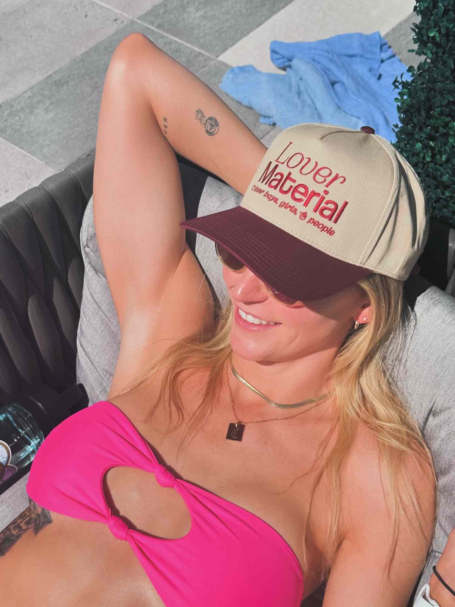 Woman wearing pink bikini top, layered necklaces, and Lover Material baseball cap