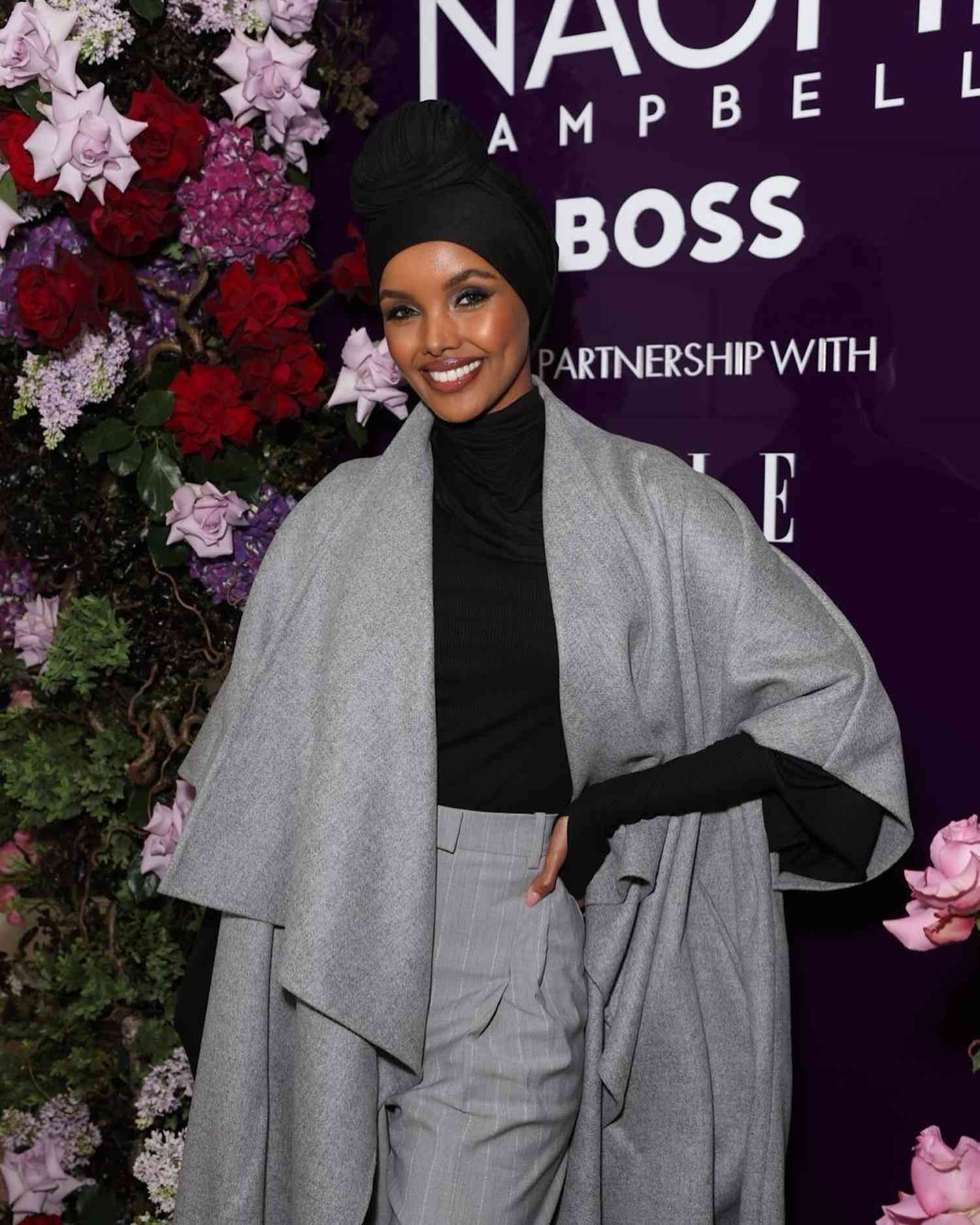 Halima Aden wears a black turtleneck, gray cape jacket, pinstriped gray pants, black head covering, and radiant makeup