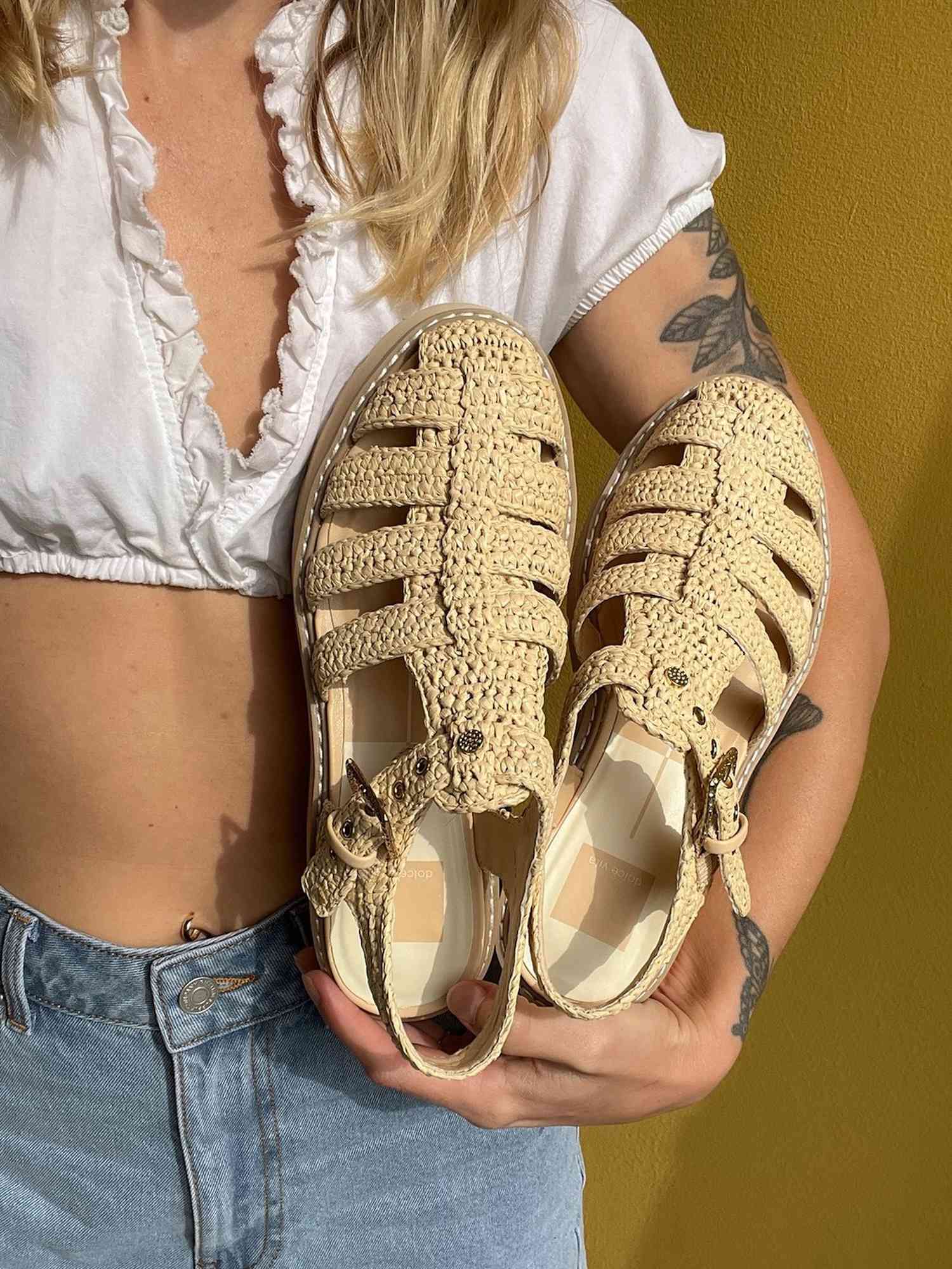 Close-up of woman holding woven fisherman sandals while wearing cropped white shirt and jeans