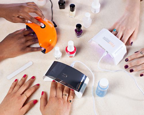 Multiple people’s hands using various nail curing lamps surrounded by different nail polishes displayed on table