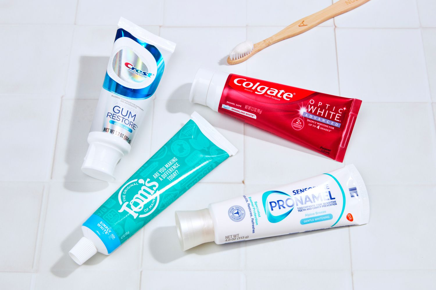 Four whitening toothpastes arranged with a toothbrush on a white tiled surface