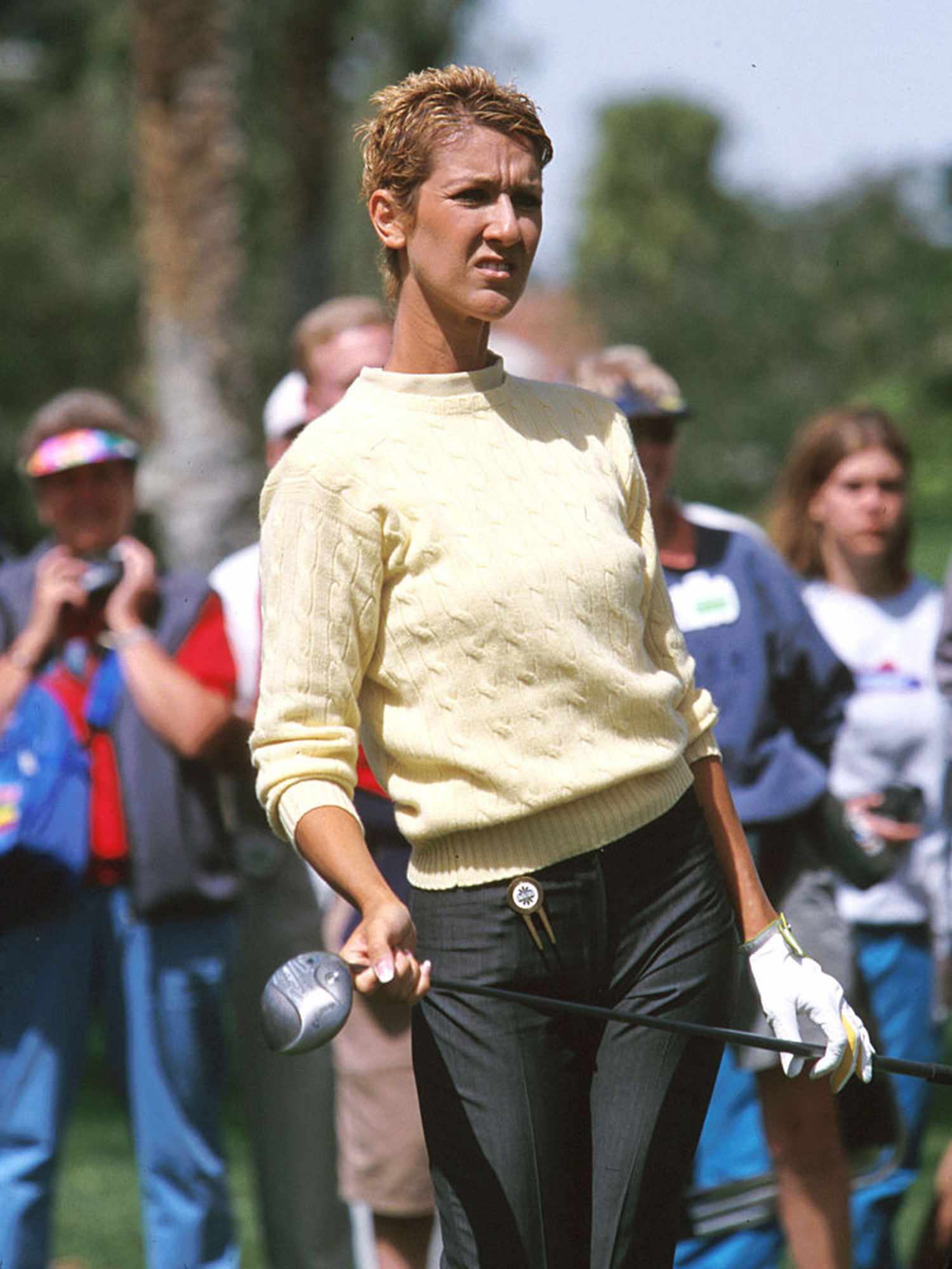 Celine Dion holding a golf club wearing yellow sweater, dark jeans, and short blonde hair cut