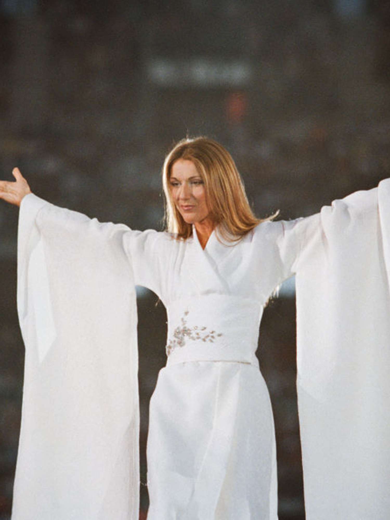 Celine Dion posingd facing the crowd with arms in the air wearing all white, kimono style outfit with dramatic sleeves