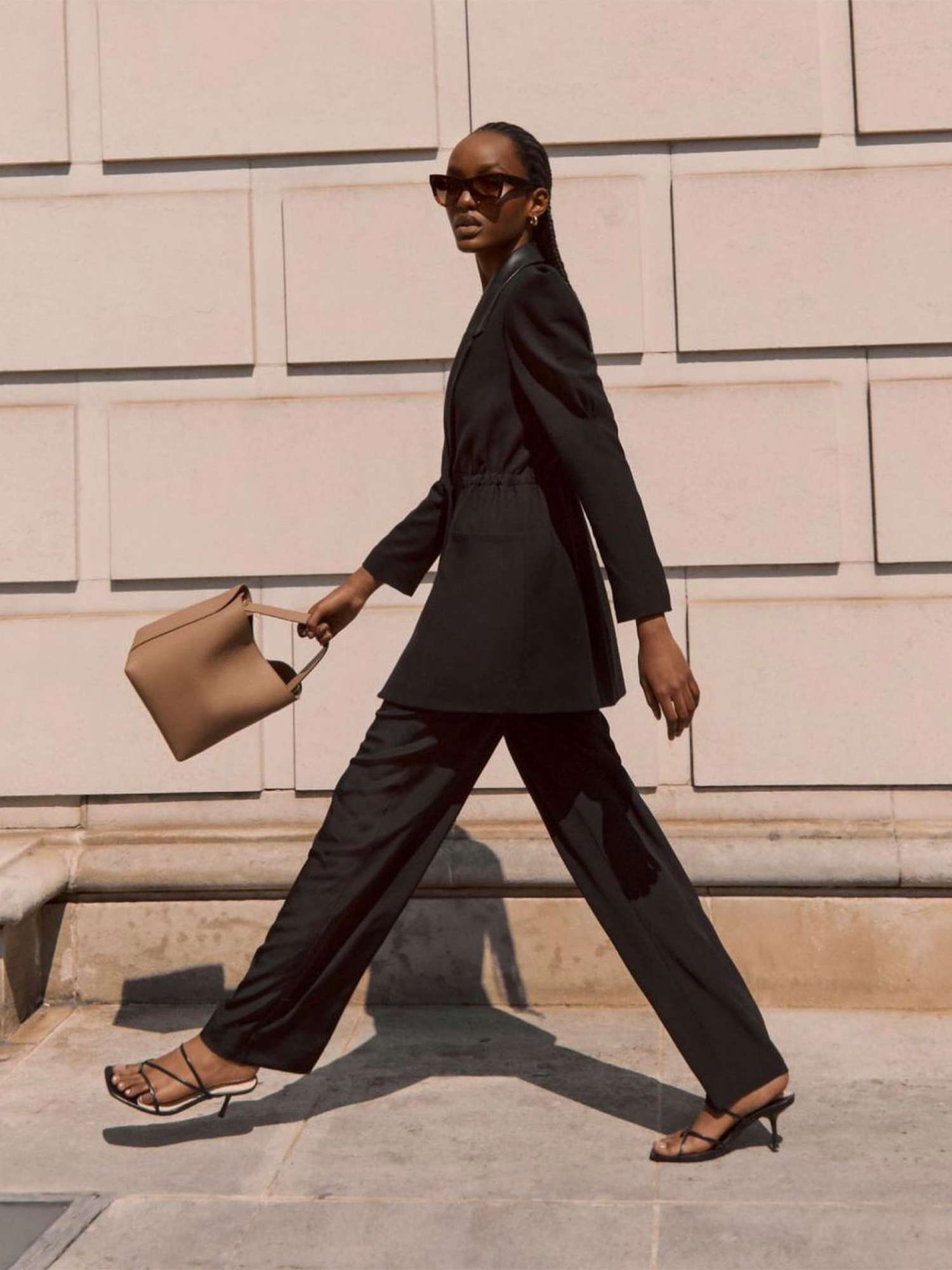 Woman carrying neutral Cuyana handbag and wearing all-black outfit while walking