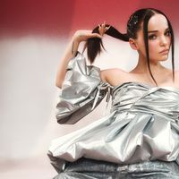 Dove Cameron holding her hair in a silver dress in front of red and white background