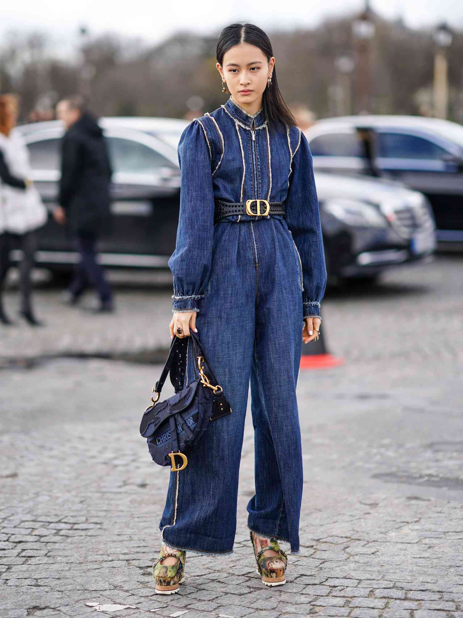 Lala Takahashi wears a blue denim jumpsuit with Dior belt, saddle bag, and platform shoes