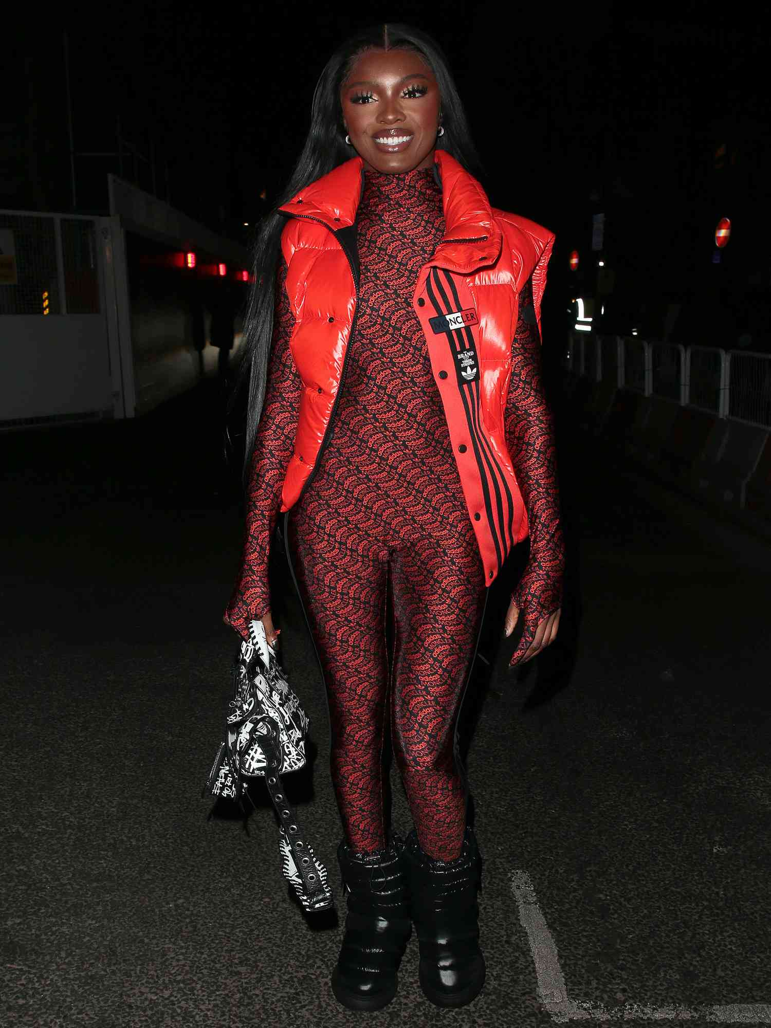 Leomie Anderson wears a red and black catsuit, red puffer vest, black and white bag, and black puffer boots. 