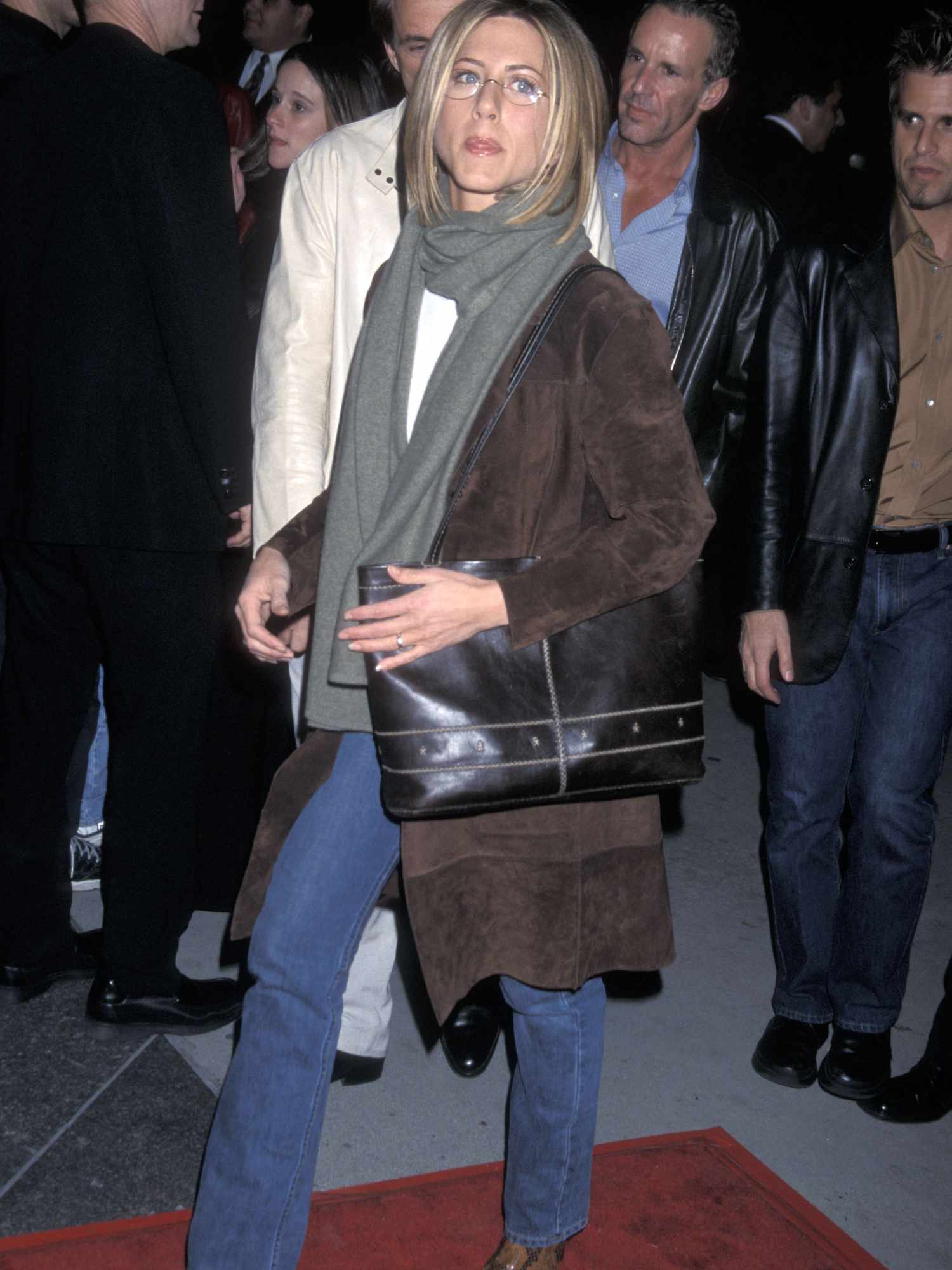 Jennifer Aniston wears a brown suede coat, gray scarf, black oversized shoulder bag, jeans, and glasses