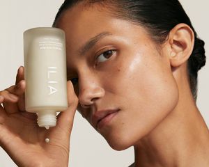 Product image of a model dropping a drop of Ilia's new Base Face Milk onto her hand