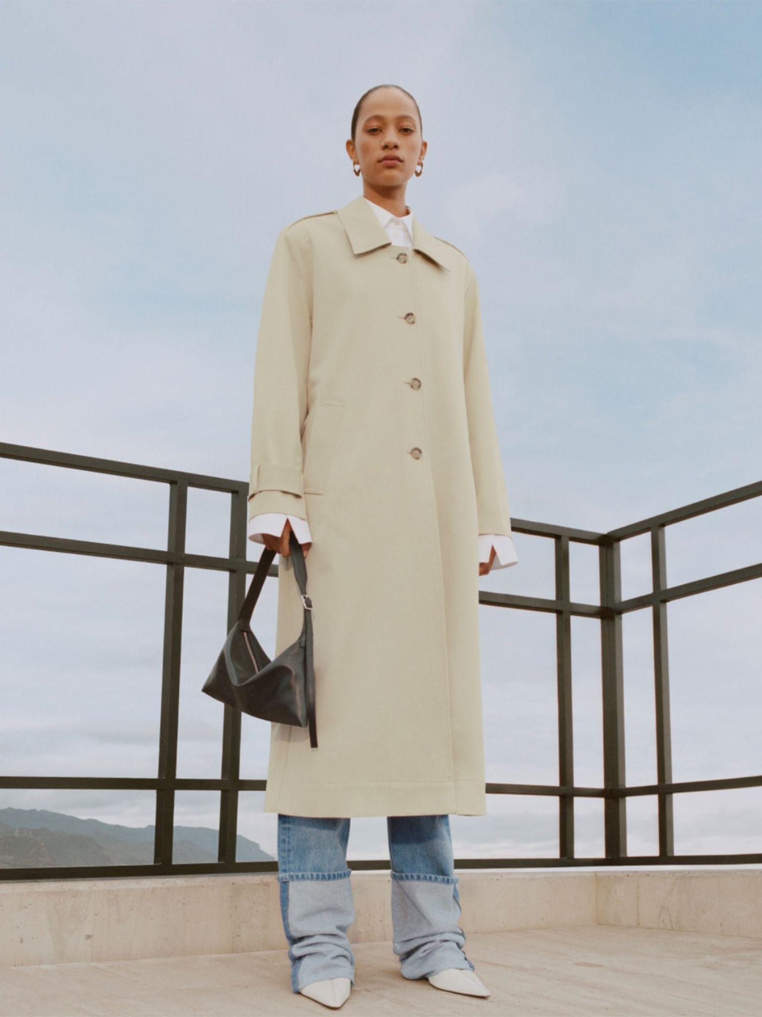 Woman wearing beige longline coat, white button-down, dark handbag, jeans, and white pointed-toe shoes from Mango