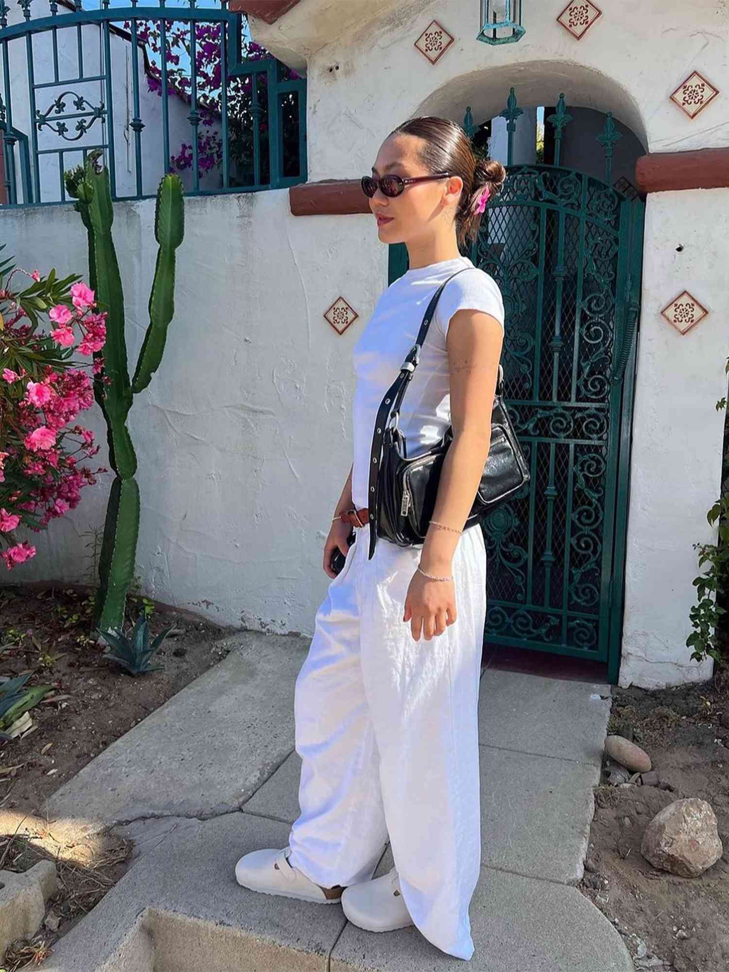 Woman carrying a black Marge Sherwood bag and wearing an all-white outfit with dark oval sunglasses and slicked-back bun hairstyle