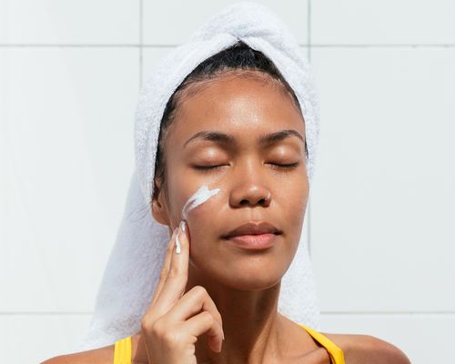 woman with hair in towel applying moisturizer to face