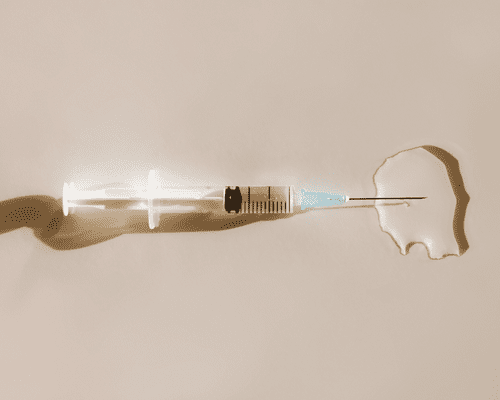 A close up of a clear syringe leaking clear liquid on a beige background.