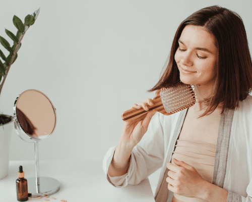 Woman brushing her hair and sitting at a vanity with an oil dropper bottle and mirror on it