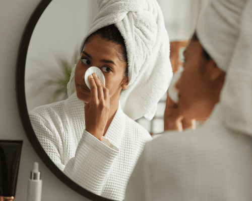 woman using cotton round to remove makeup in mirror