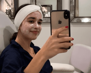 A woman with numbing cream and a headband in a doctor's office taking a mirror selfie