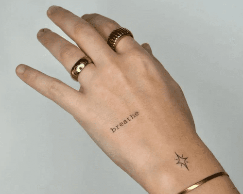 A hand wearing gold wrings and a bracelet with a "breathe" tattoo on top and a starburst tattoo on the wrist